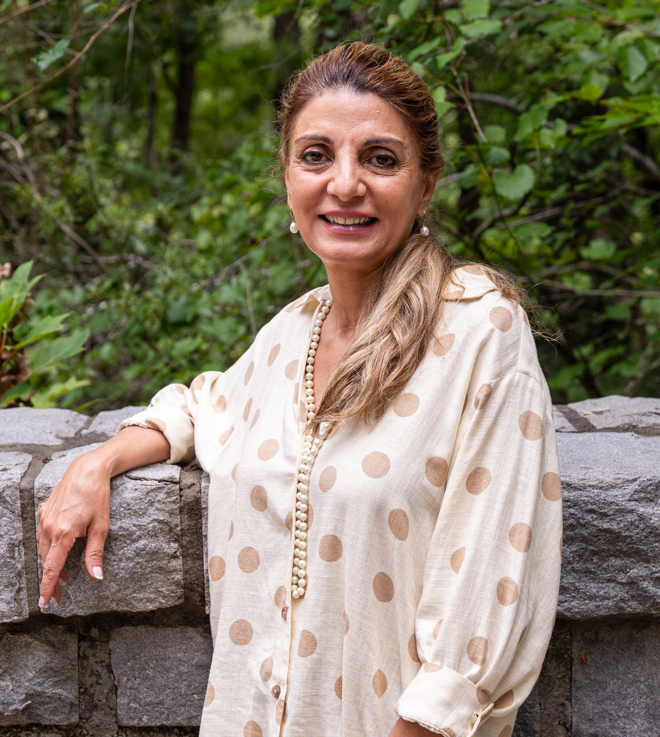 Woman with long brown hair stands outdoors by a stone wall, wearing a cream shirt with large polka dots and a beaded necklace, with green foliage in the background.