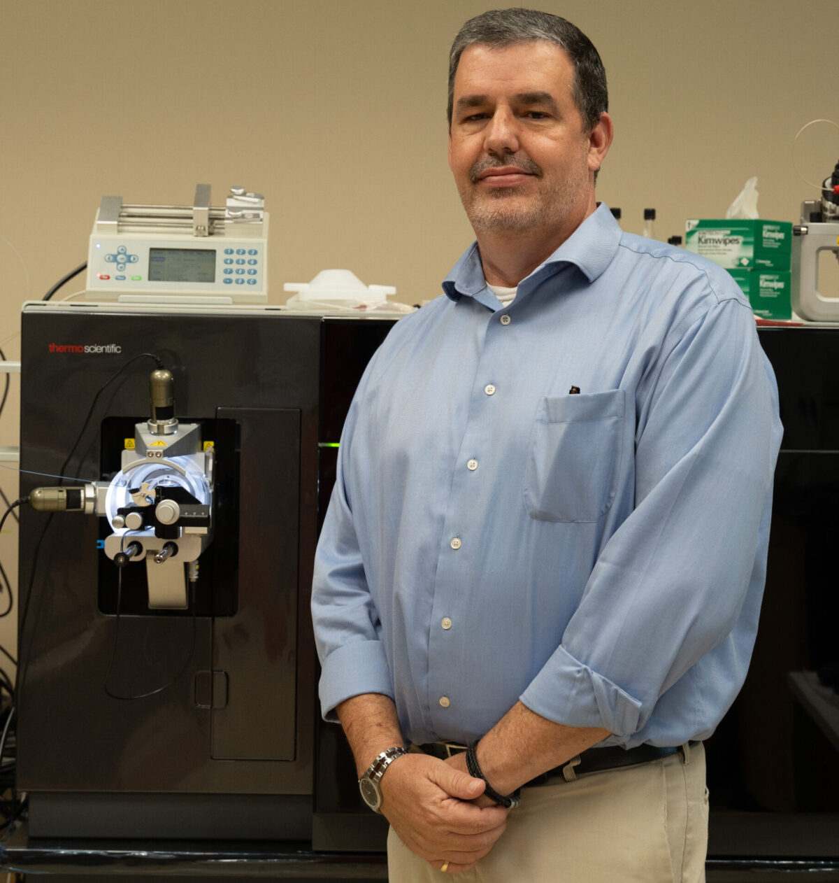 A man in a blue shirt stands next to a Thermo Scientific laboratory instrument in a research lab setting.