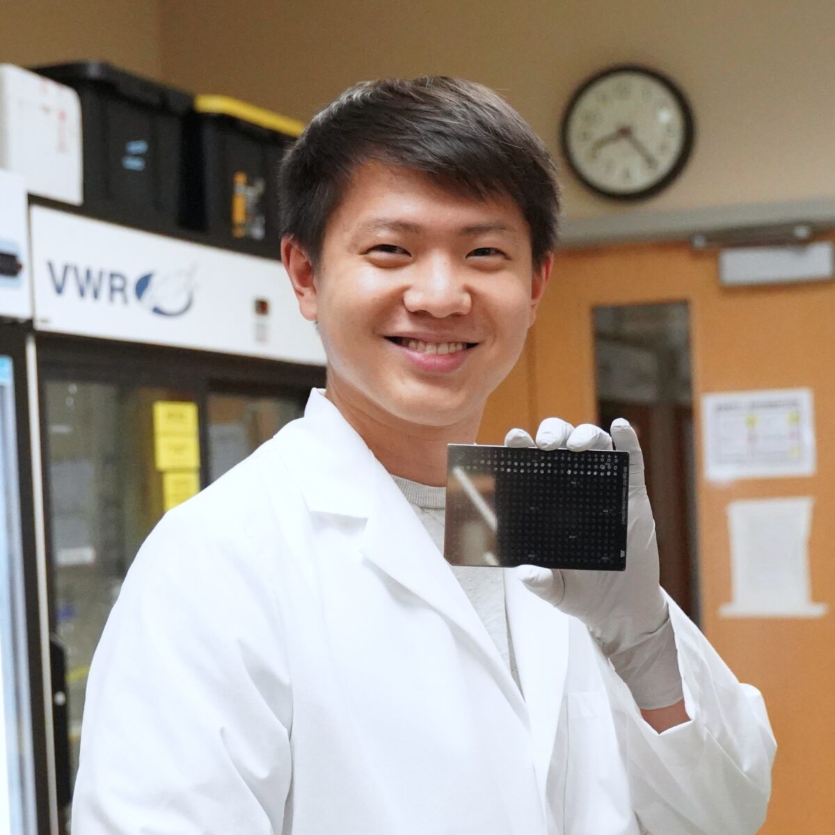 Person in a white lab coat and gloves holds up a rectangular scientific plate or grid in a laboratory setting with a clock in the background.