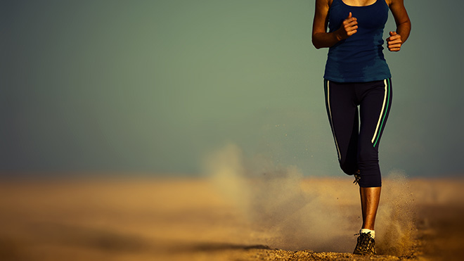 A runner in a blue tank top and athletic leggings jogging across sand, kicking up dust