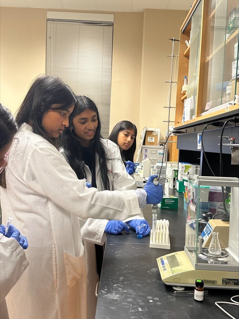 Three people wearing lab coats and blue gloves work with pipettes and test tubes in a laboratory setting, standing at a black counter with scientific equipment.