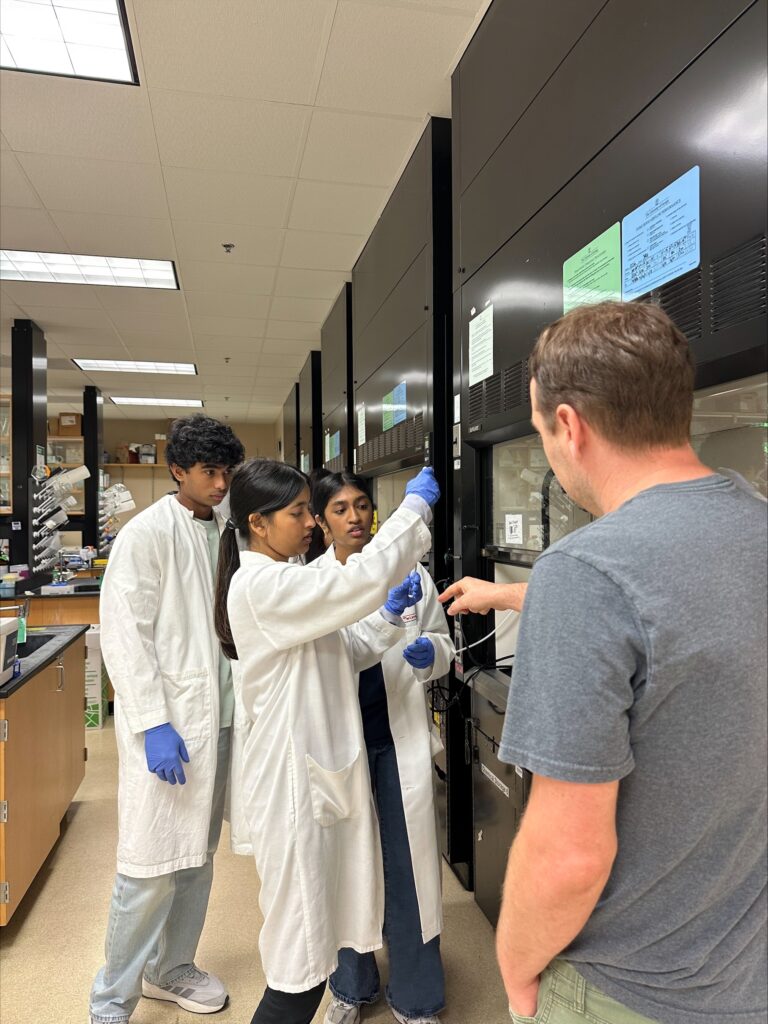 Three people in lab coats and gloves work with lab equipment while a man in casual clothes observes and gestures in a laboratory setting.