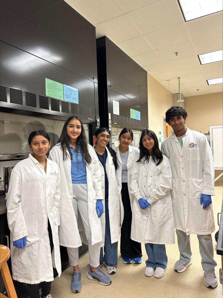 Six people wearing white lab coats stand together in a laboratory, some wearing blue gloves. Laboratory equipment and information sheets are visible in the background.