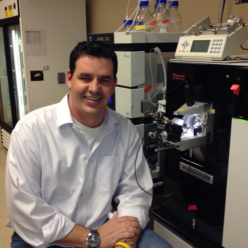 Dr. Lance Wells seated beside a Thermo Scientific mass spectrometer and UltiMate 3000 chromatography system in his laboratory at the CCRC.