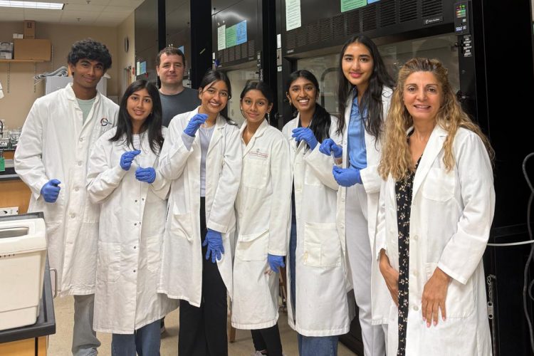 A group of seven people in lab coats and blue gloves stand together in a laboratory, posing for a photo. Laboratory equi