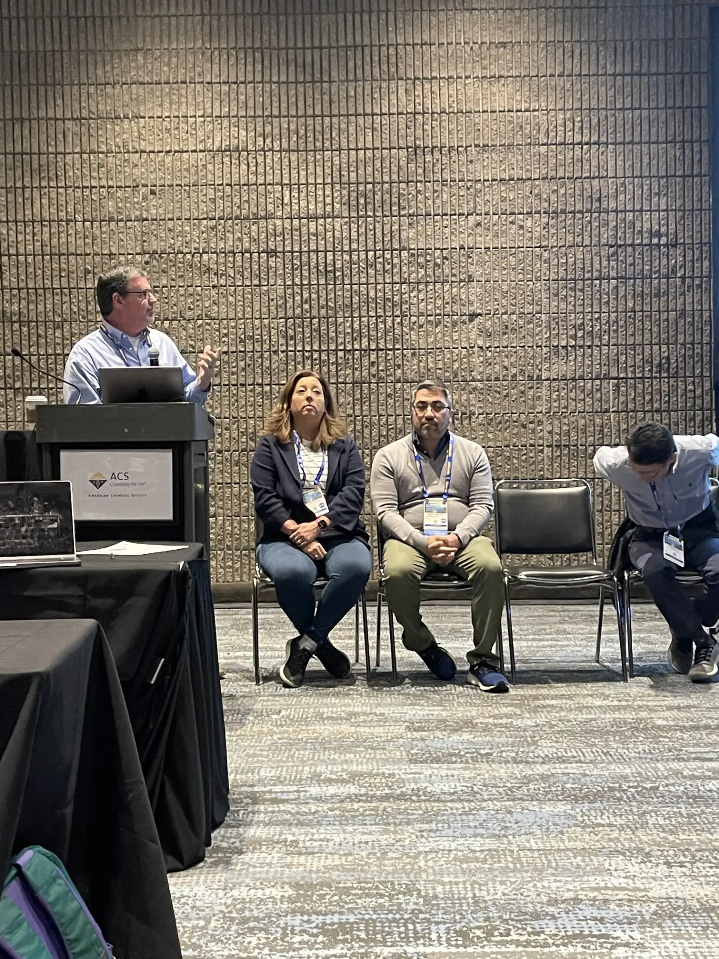 A speaker at a lectern introducing three seated panellists during an ACS session.