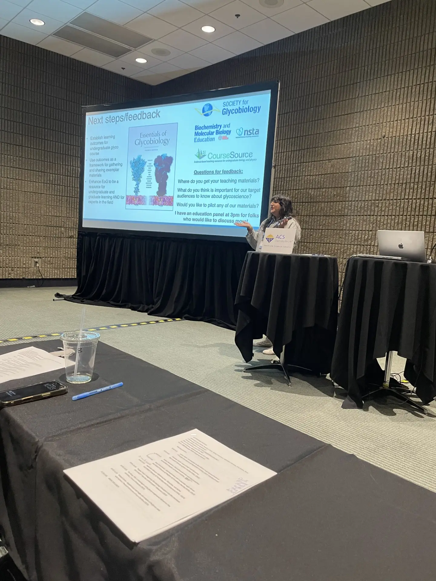 A speaker presenting a slide headed "Next steps/feedback" on glycoscience teaching materials and learning outcomes.
